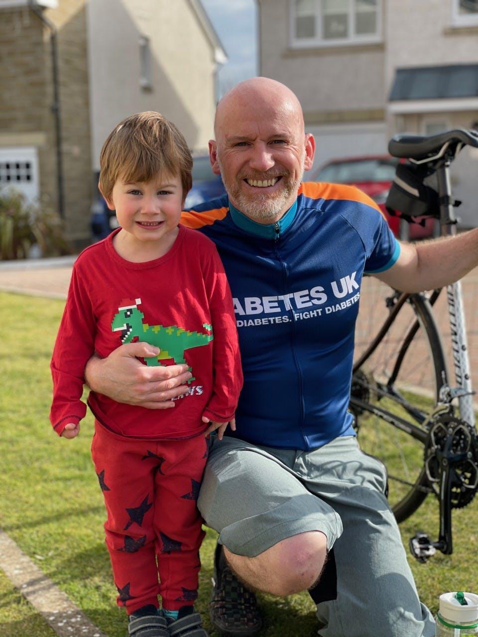 Euan in Diabetes UK cycling jersey holding his bike with his other arm around his son Archie in red pyjamas